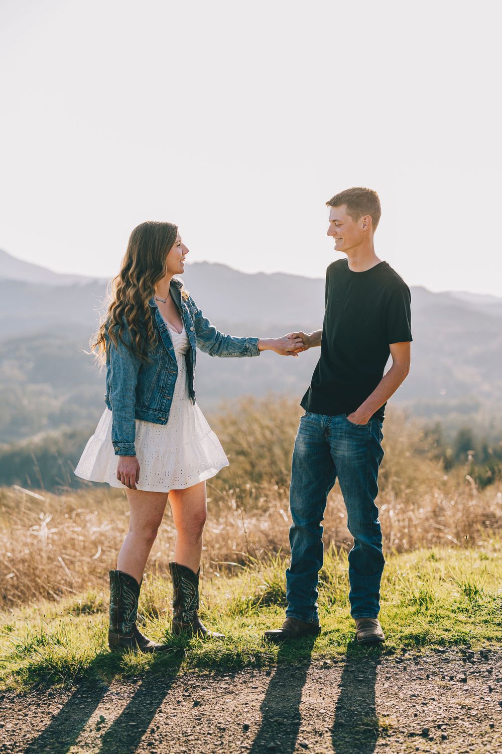 A couple stands together on a hilltop during golden hour wearing casual outfits against a mountain backdrop.