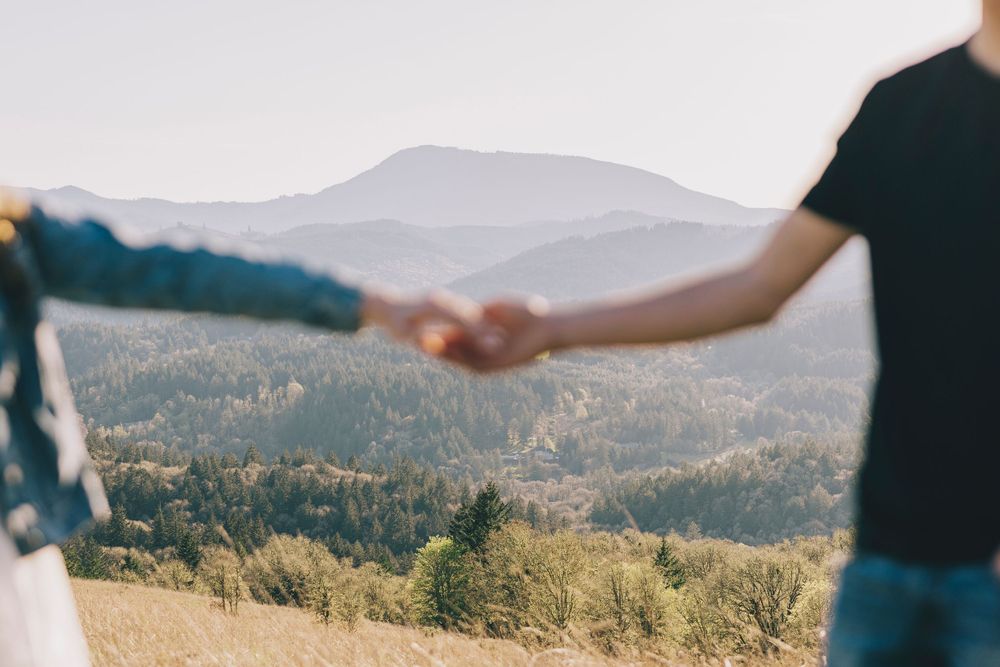 A couple holds hands against a scenic mountain landscape with rolling hills and evergreen forests in the background.