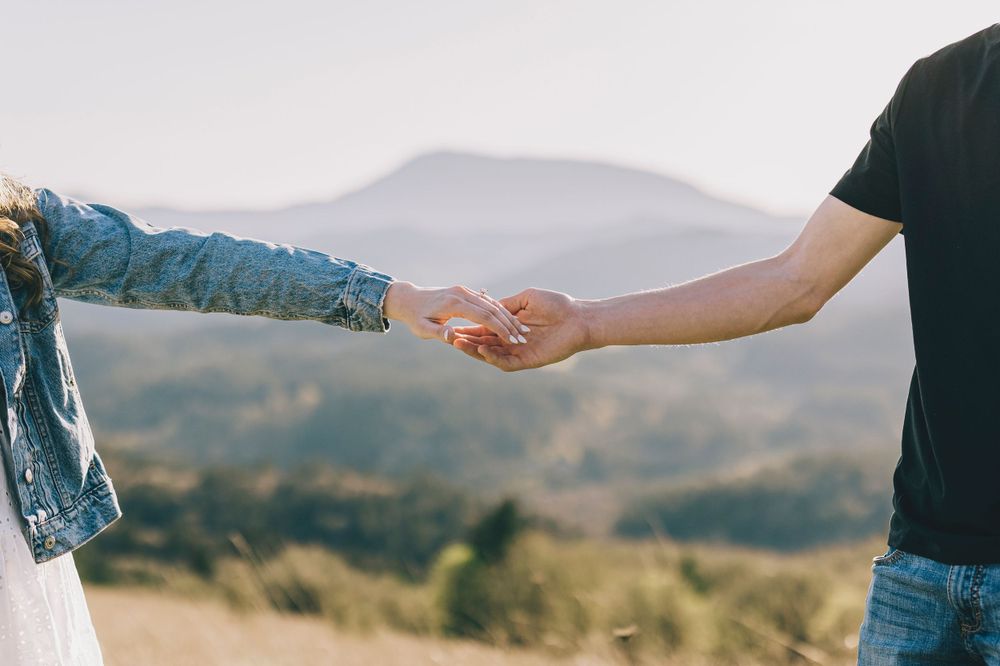 Two hands reaching out to hold each other against a scenic mountain landscape backdrop.