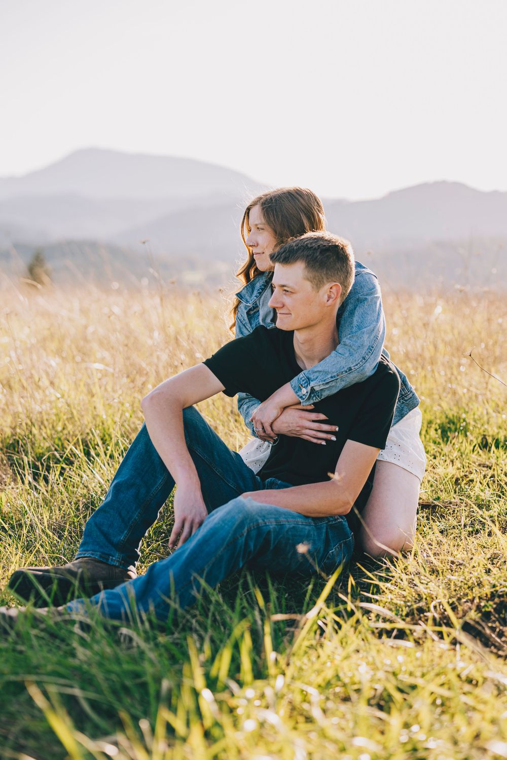 A couple shares a tender moment sitting together in a golden field with mountains in the background at sunset.