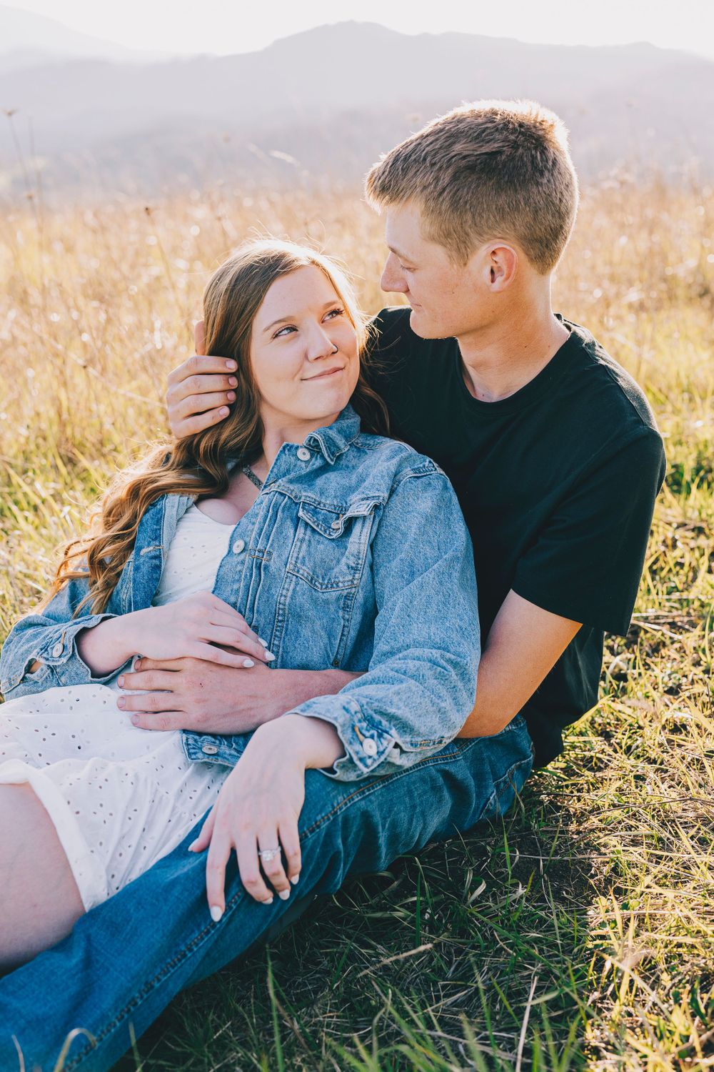 A loving couple sits together in a golden field at sunset with mountains in the background.