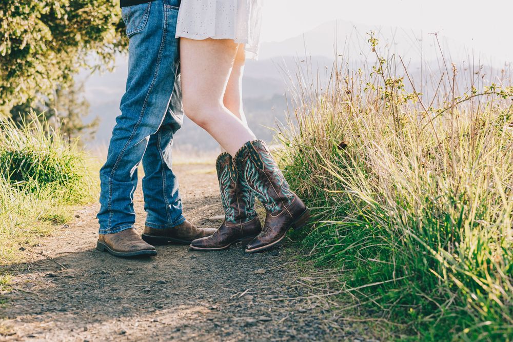 Couple's feet in boots standing close together on a dirt path surrounded by tall grass with mountains in the background.