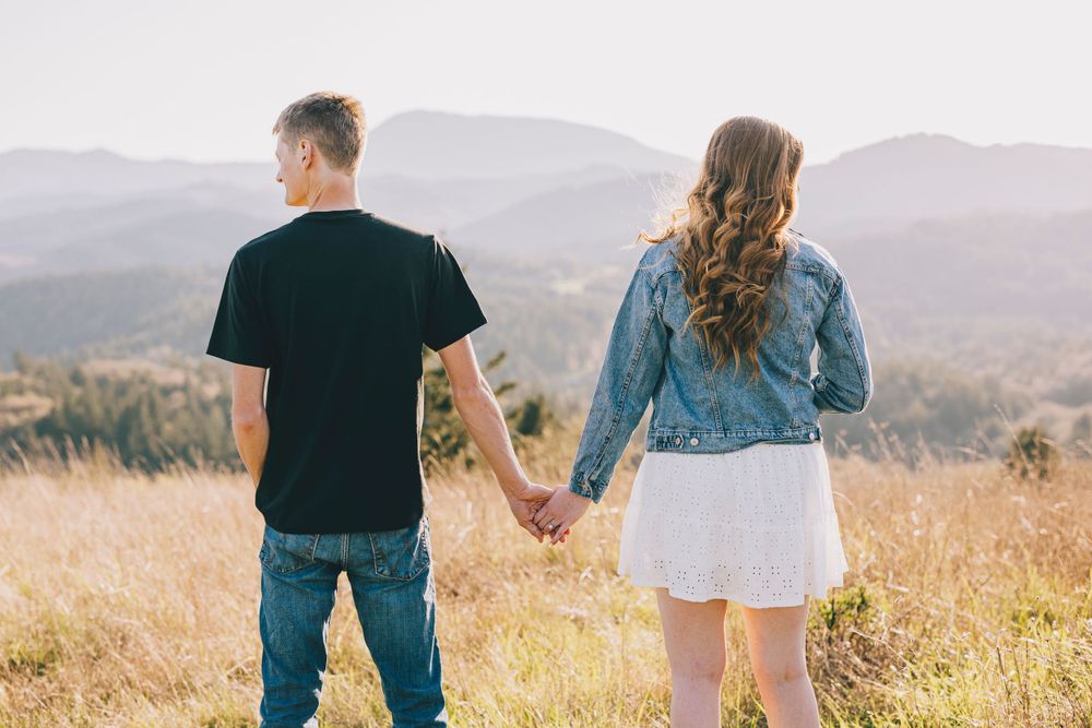 A couple holding hands while standing in a golden field with mountain views in the background.