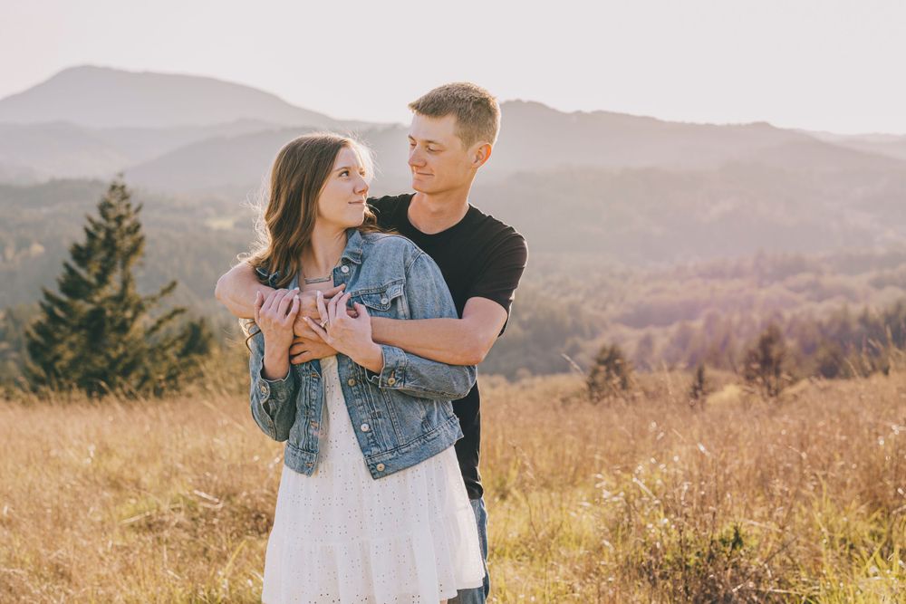 Two people embracing in casual attire on a hilltop overlooking mountains during sunset.