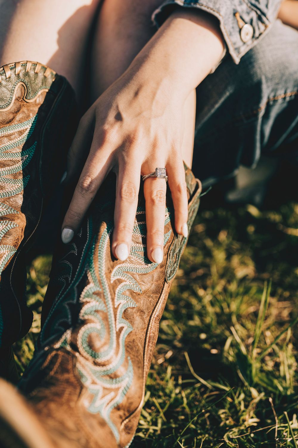 A hand rests gently on a brown leather cowboy boot with intricate western stitching in sunlit grass.