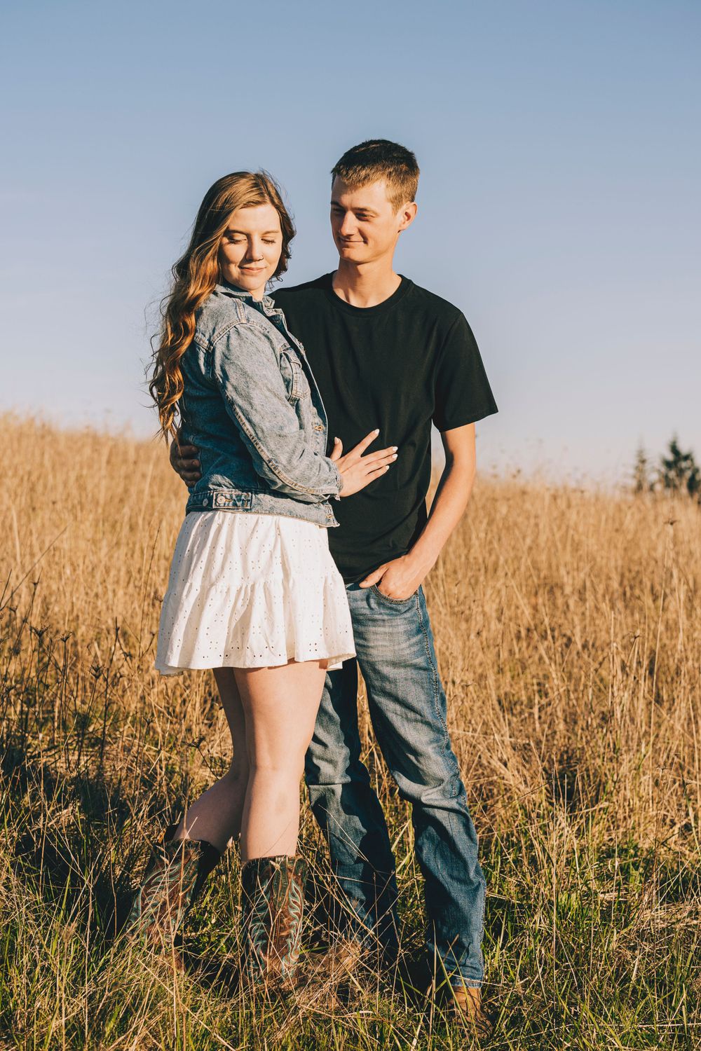 A couple in casual attire poses together in a golden field with mountains in the background.