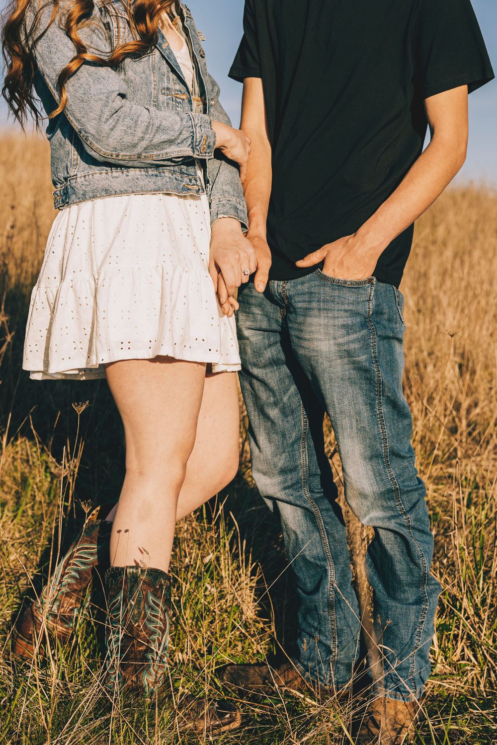Couple standing in golden field wearing denim and lace outfit with boots in warm sunset lighting.