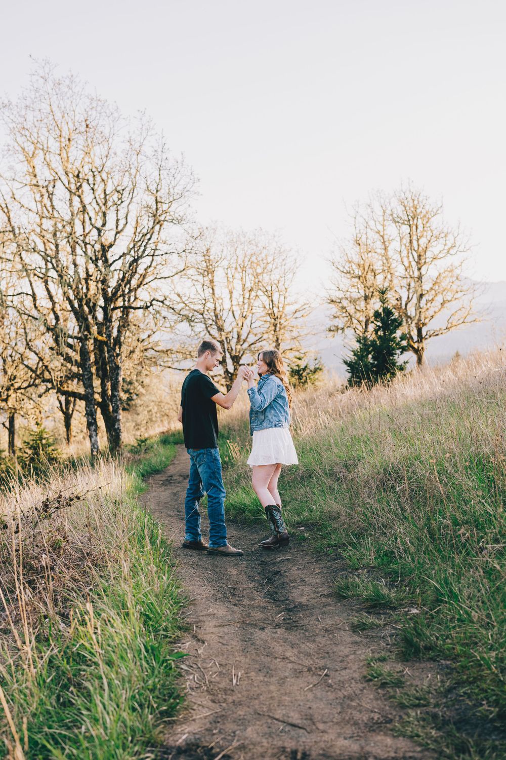 Two people standing together on a dirt hiking trail surrounded by barren trees during sunset.