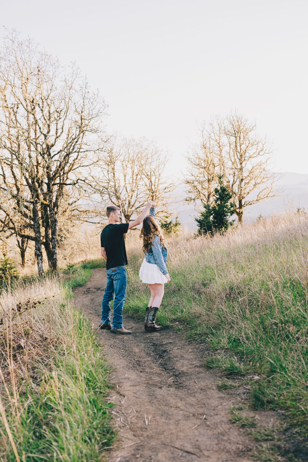 Two people walk hand in hand along a dirt trail surrounded by bare trees and autumn grass.