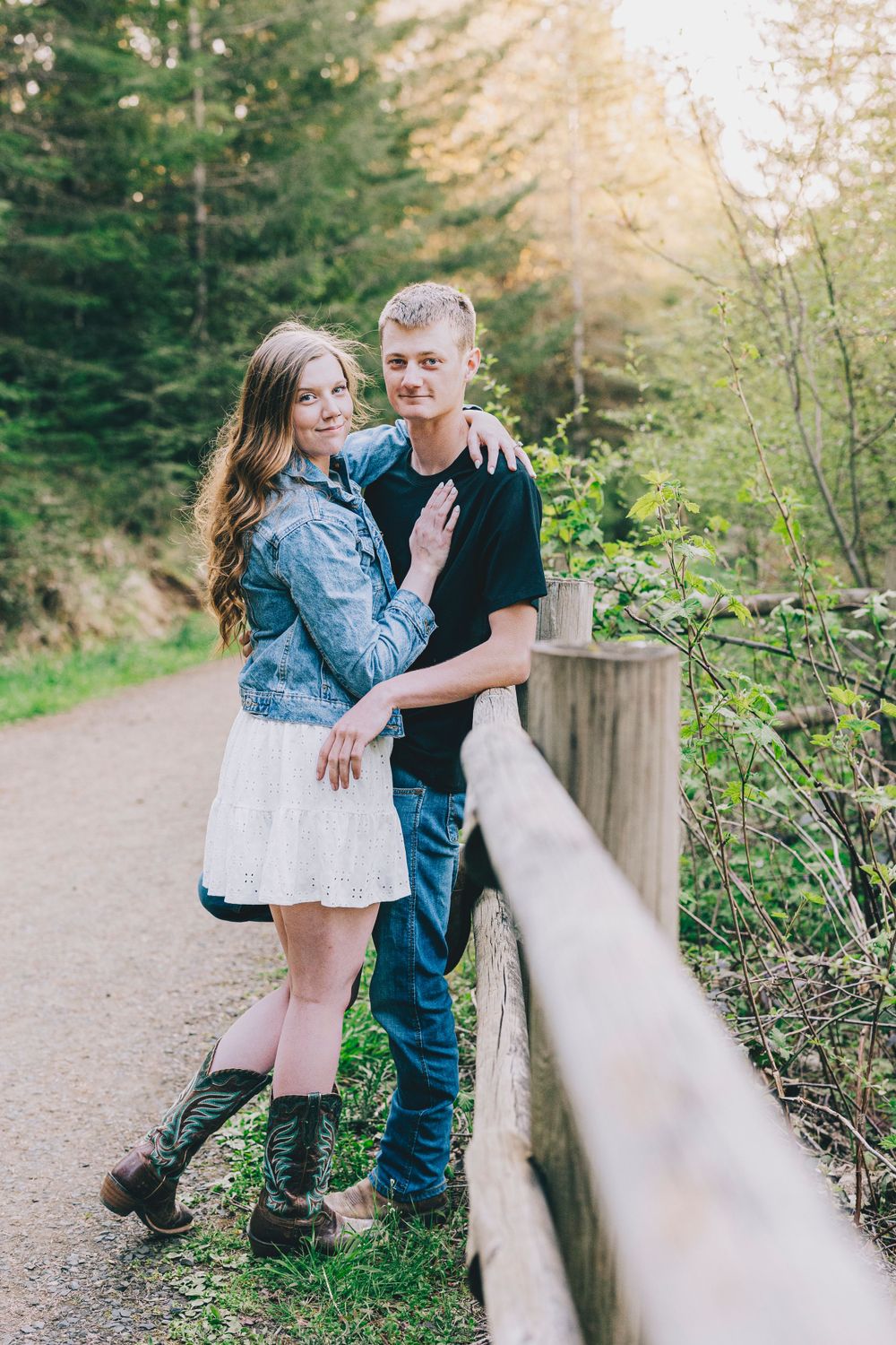 A couple poses together on a woodland path beside a wooden fence in spring.