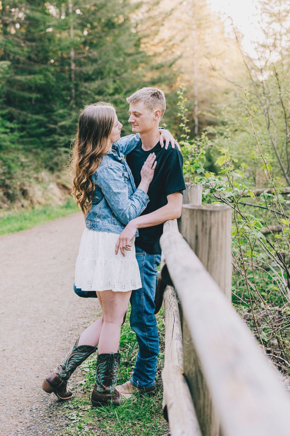 A couple embraces by a wooden railing on a forest path with evergreen trees in the background.