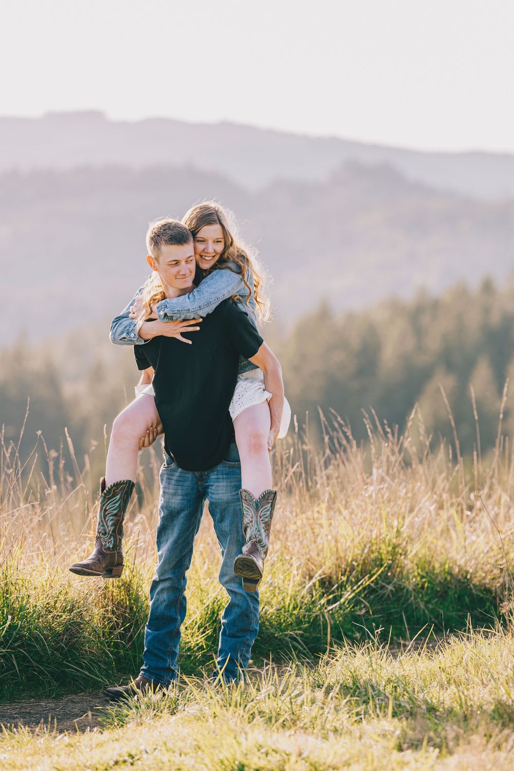 A happy couple sharing a playful piggyback ride moment in a sunlit mountain meadow with tall grass.