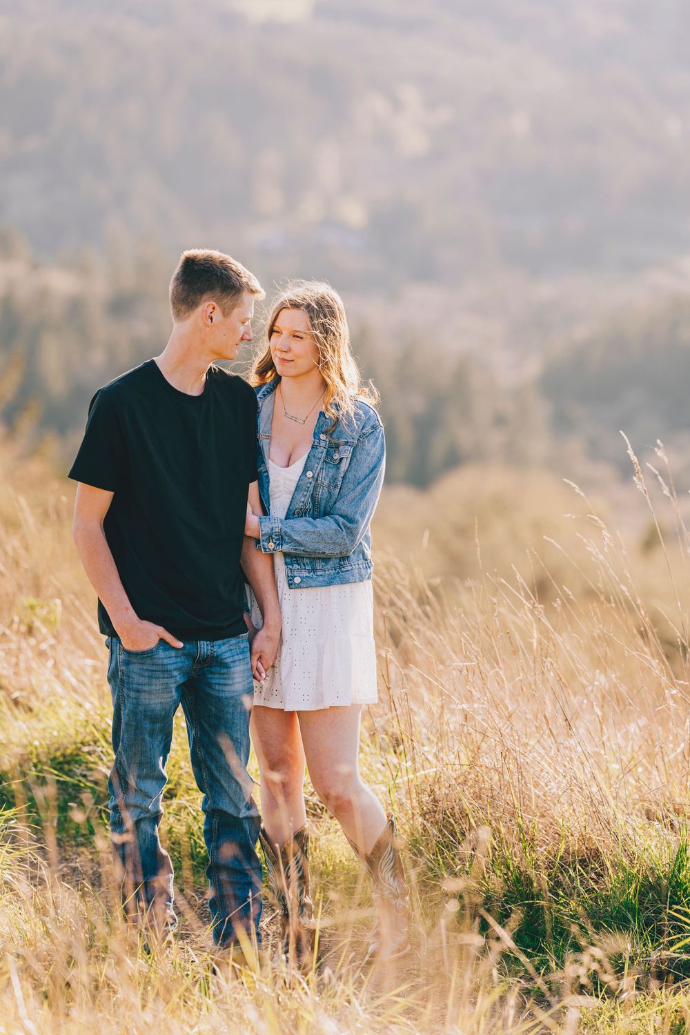 A romantic couple stands together in golden mountain meadow wearing casual attire at sunset.