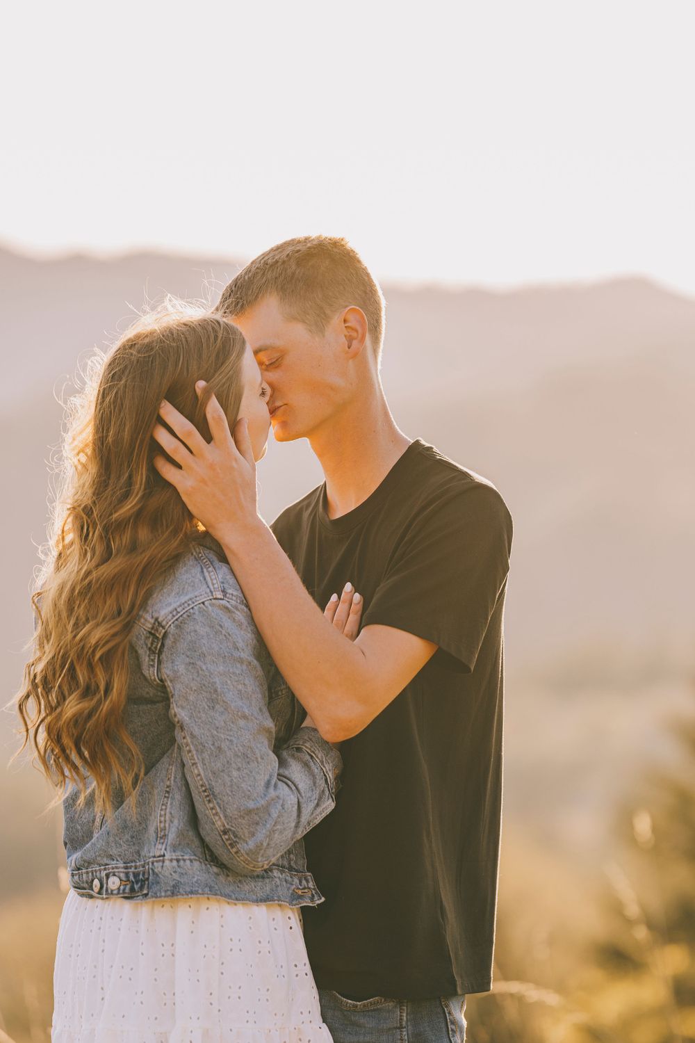 A romantic couple shares an intimate moment in warm sunlight with mountains visible behind them.