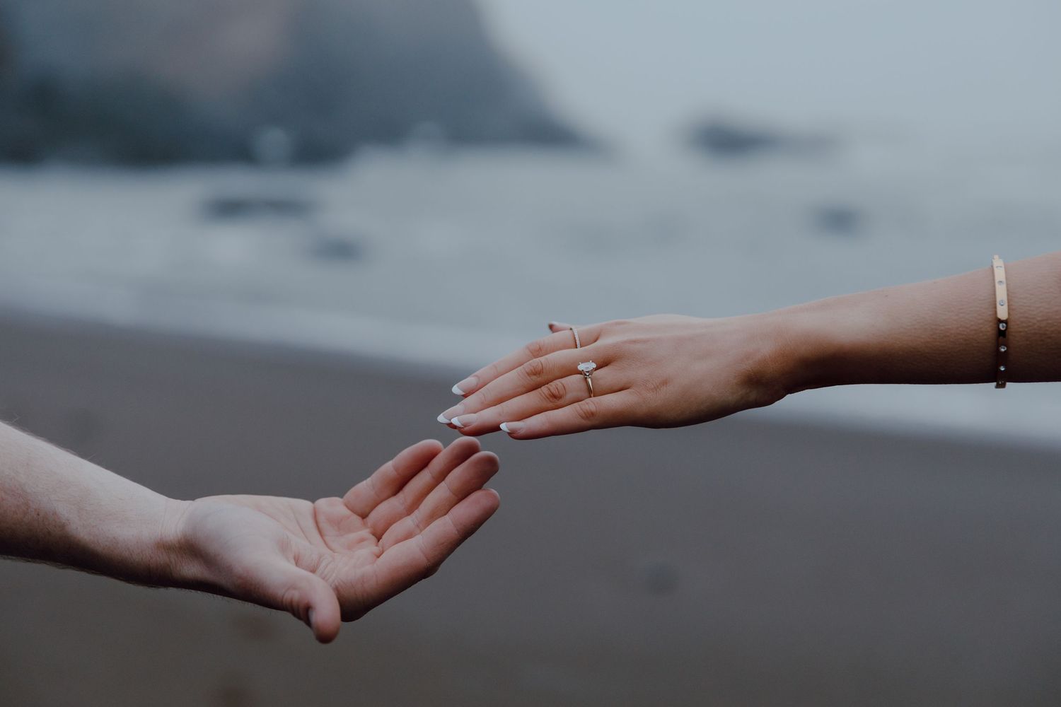 Two hands reaching toward each other on a misty beach, one hand wearing a delicate bracelet.