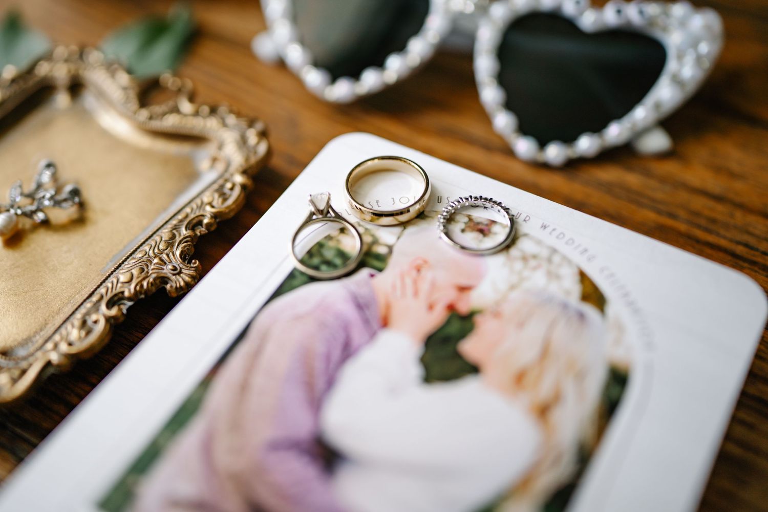 Wedding rings rest on a photograph alongside decorative heart-shaped sunglasses and vintage mirror on wooden surface.