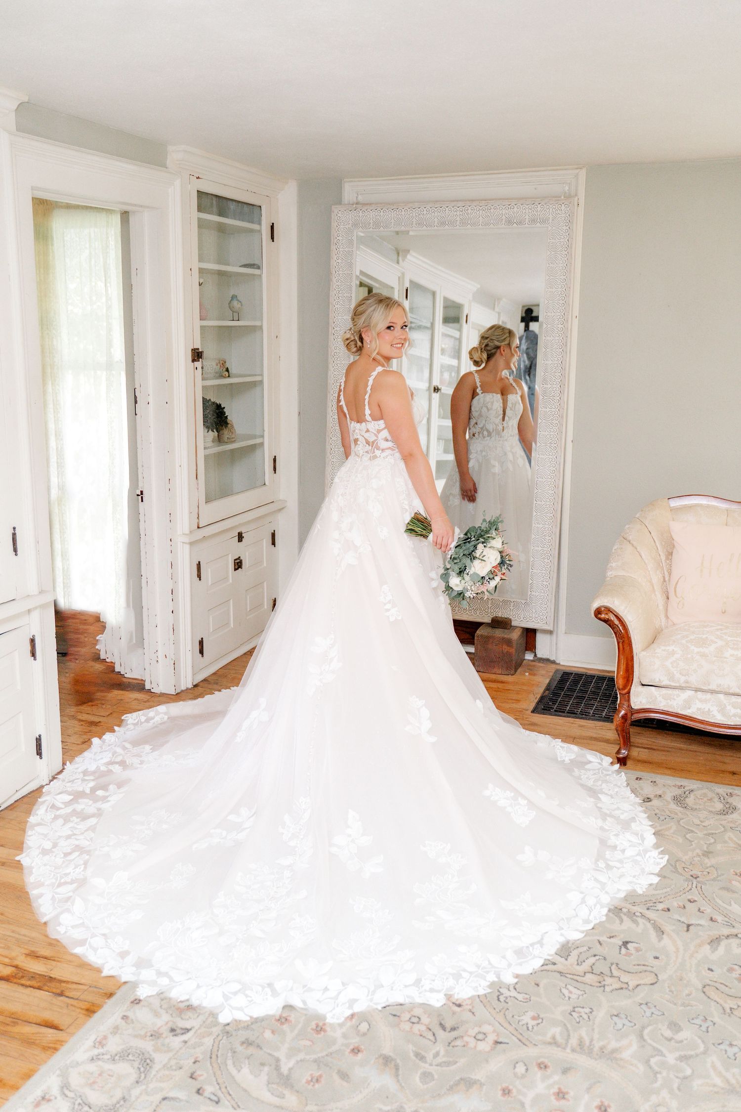 A bride in an elegant white lace wedding dress with long train poses in front of a full-length mirror in a bright room.