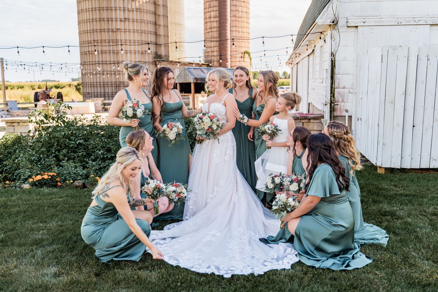A bridal party in teal dresses poses together with bouquets in front of a rustic barn setting at sunset.