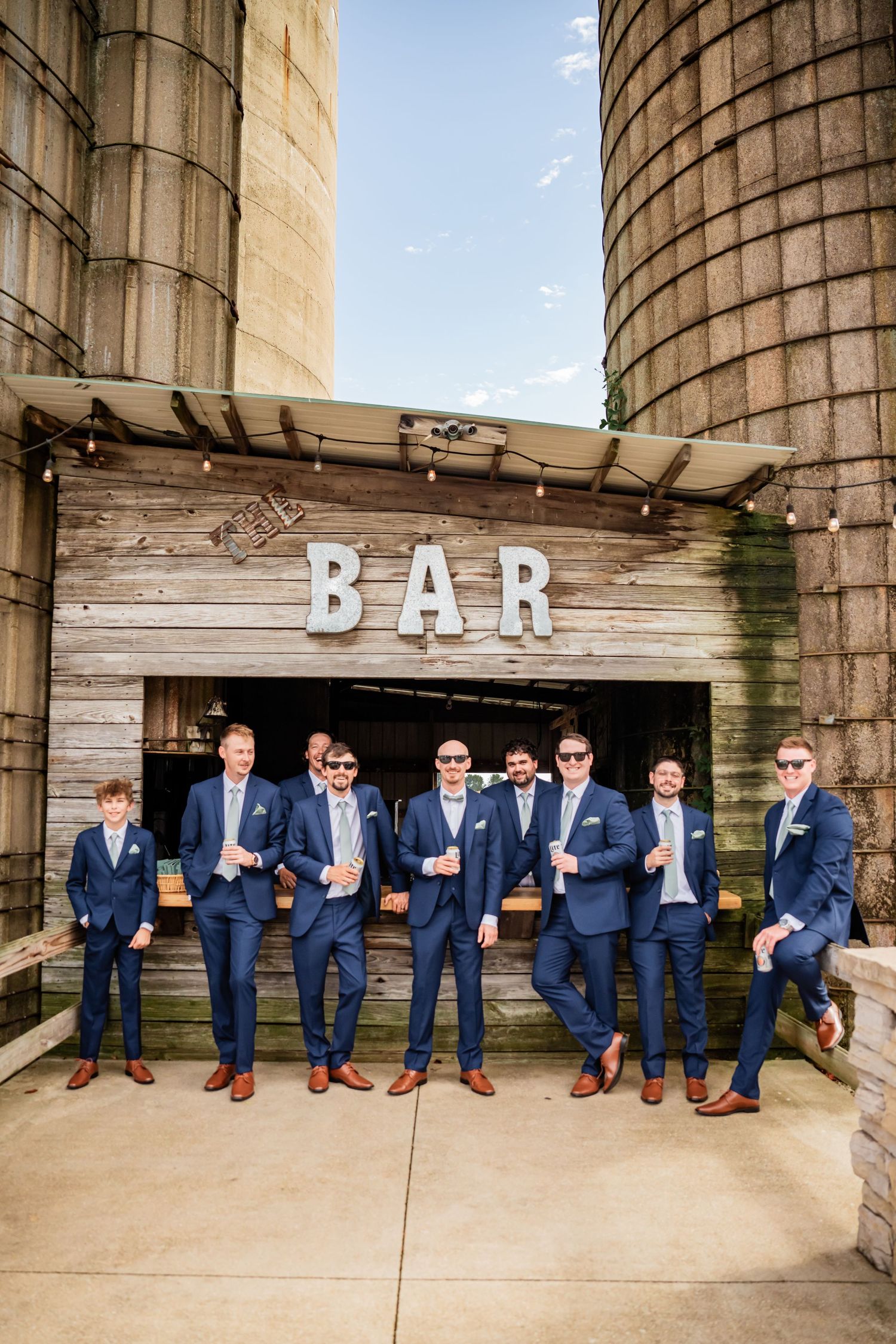 A group of groomsmen wearing matching navy suits pose together in front of a rustic wooden bar sign at a wedding venue.