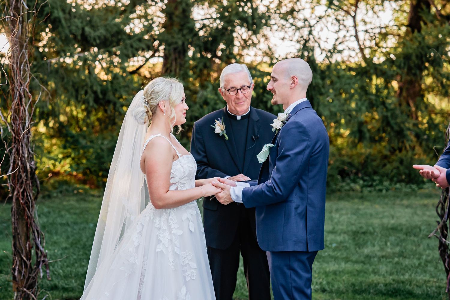 A wedding ceremony takes place outdoors on a green lawn with trees in the background during golden hour.