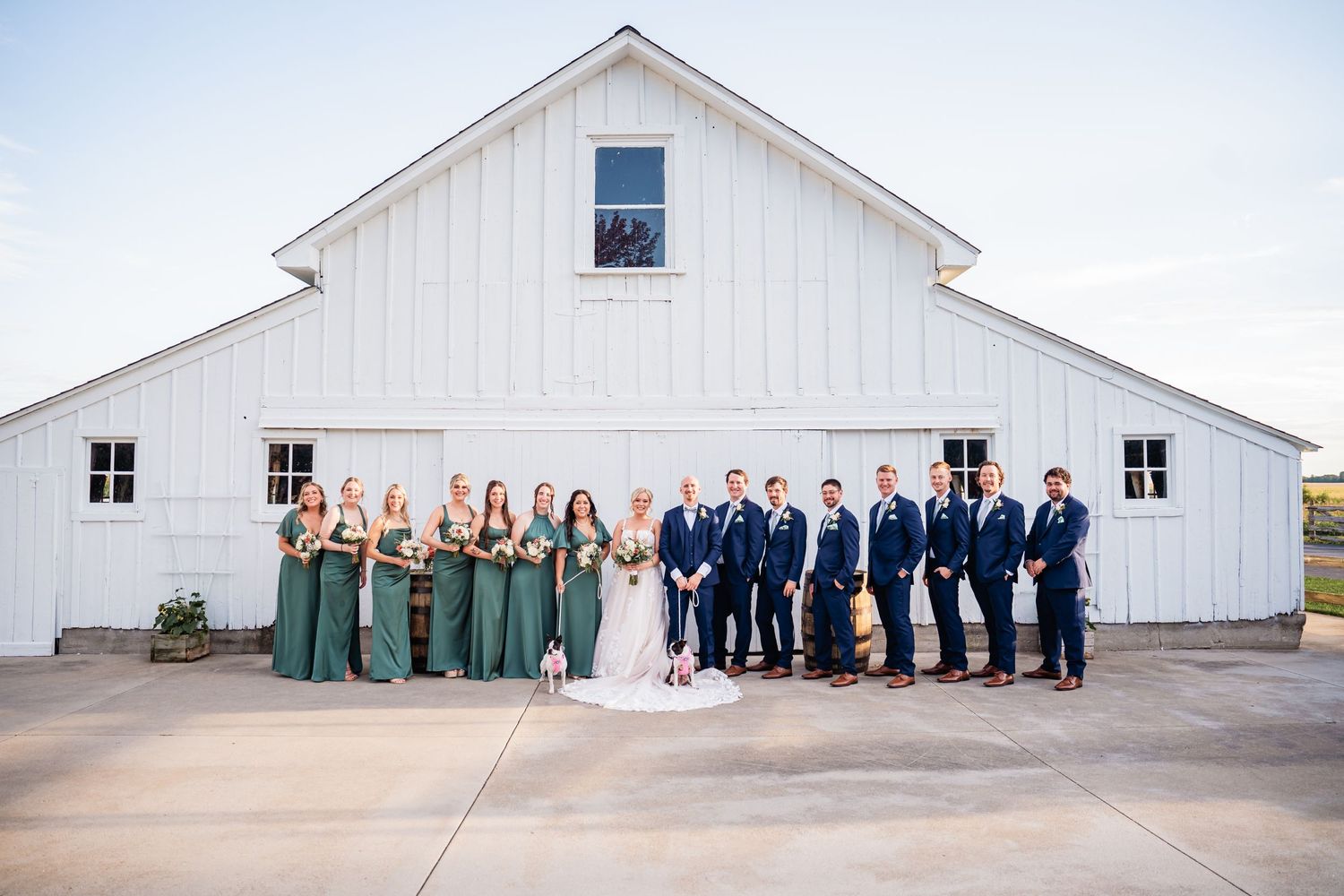 Wedding party lined up against white barn exterior with bridesmaids in green dresses and groomsmen in navy suits.
