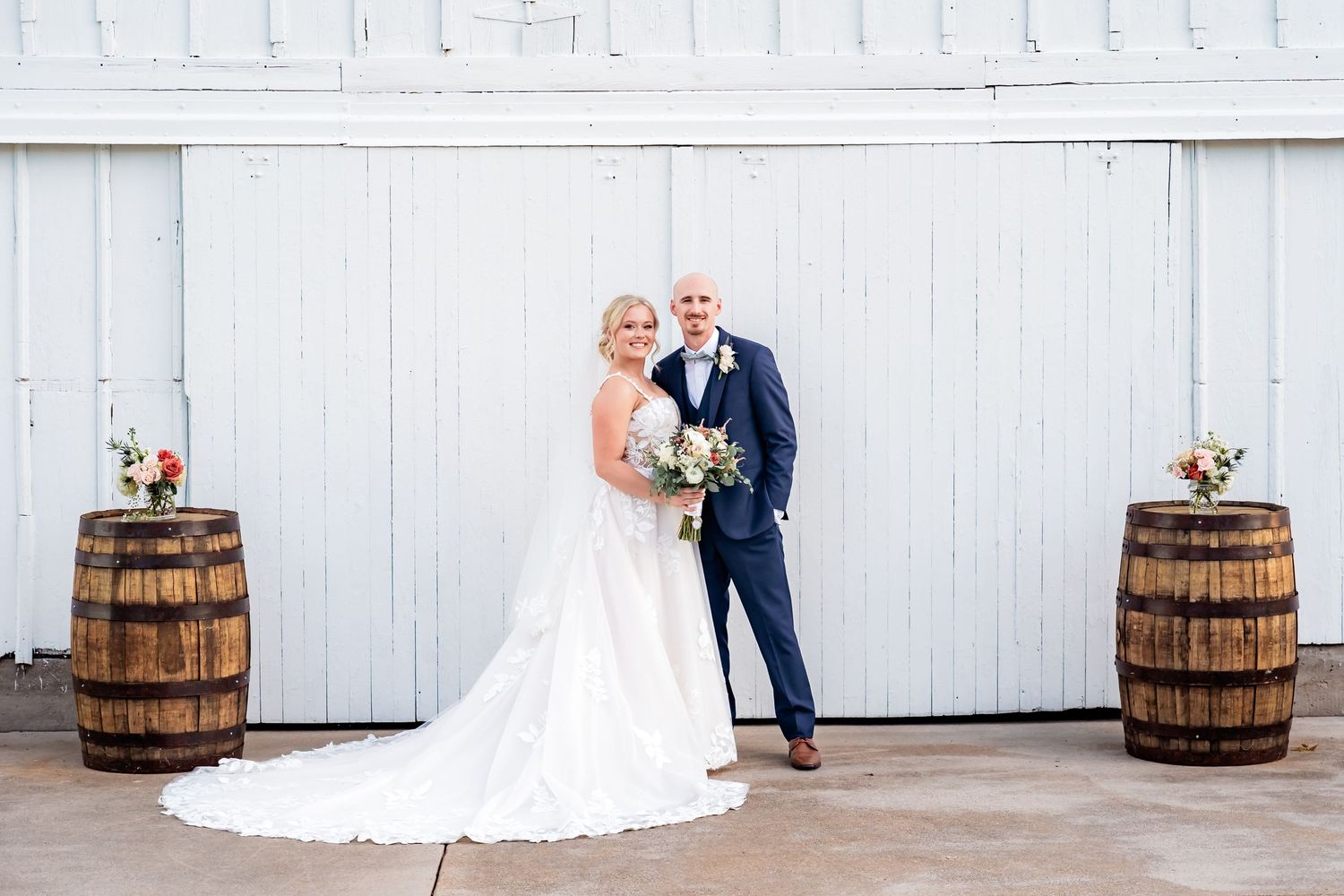 A couple poses on their wedding day against a white barn door with rustic wooden barrels and floral arrangements.