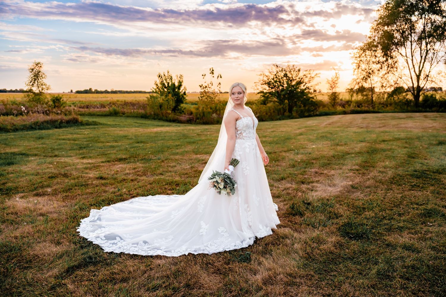 A beautiful wedding dress with long lace train photographed in a field at sunset with golden light and cloudy sky.
