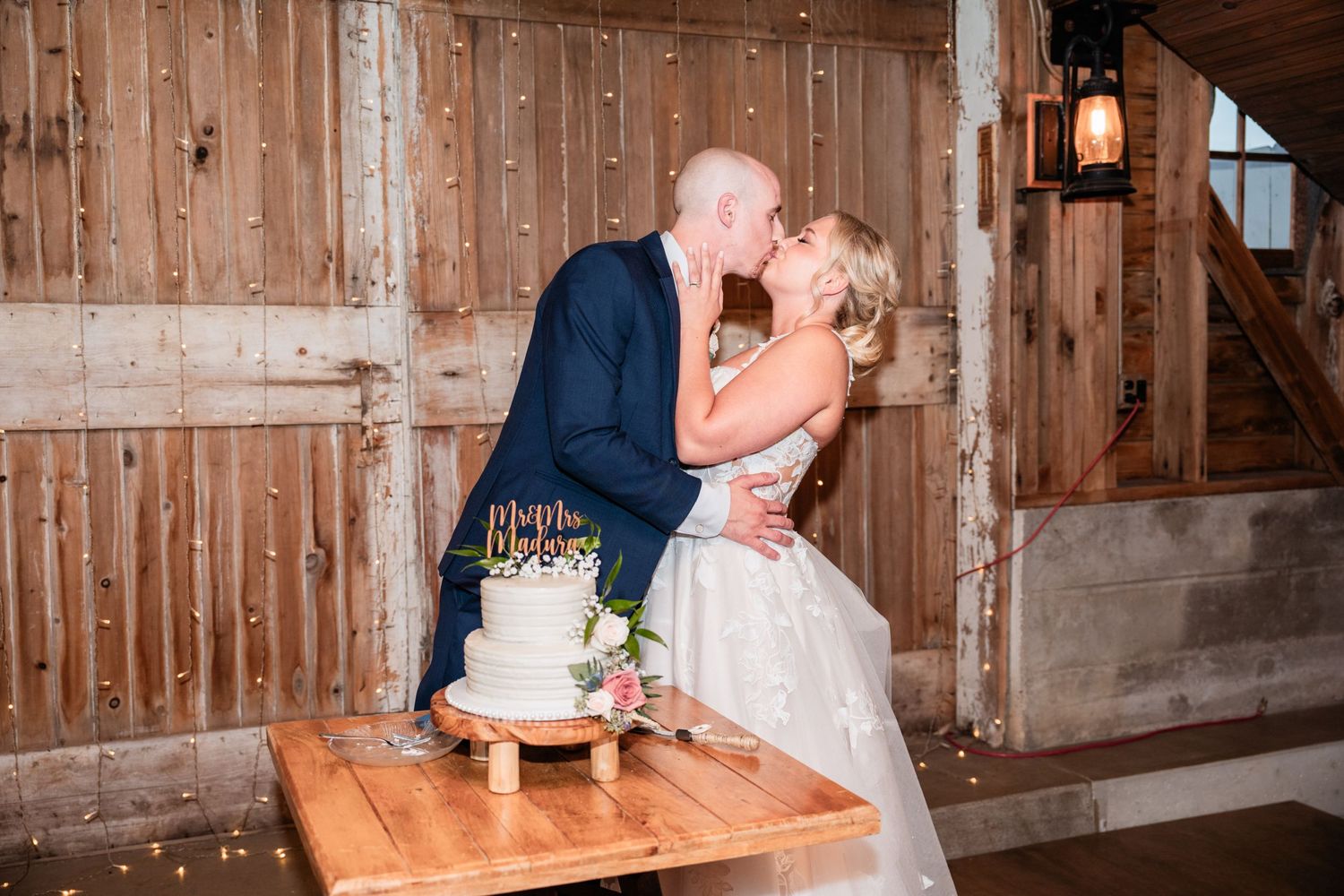 A newlywed couple kiss next to their wedding cake in a rustic wooden barn setting.