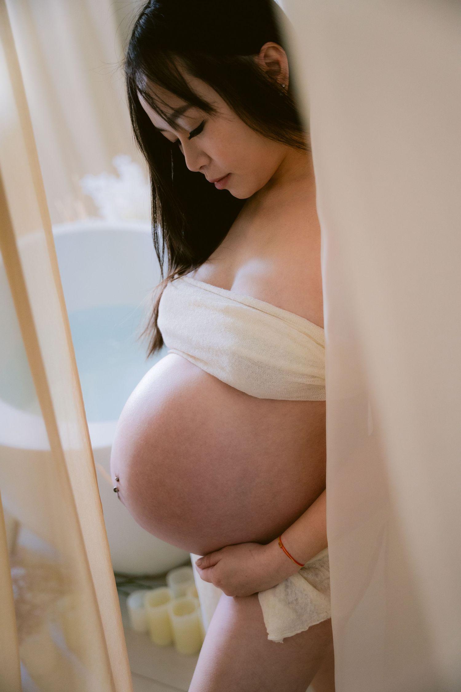 A pregnant woman in white fabric stands by a window in soft natural light during an intimate maternity photoshoot.