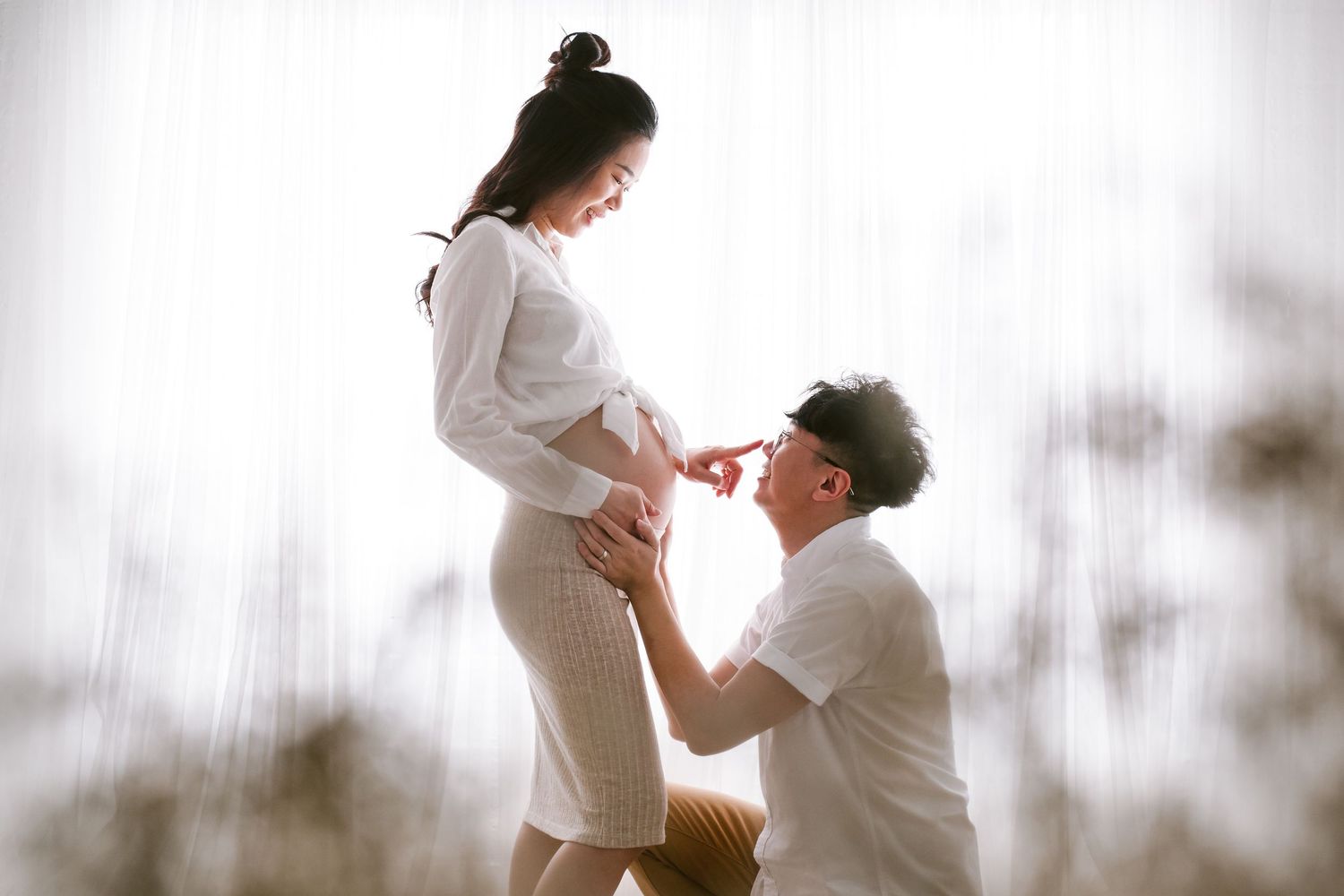 Romantic outdoor maternity portrait in soft natural light with a tender moment between a couple in white outfits.