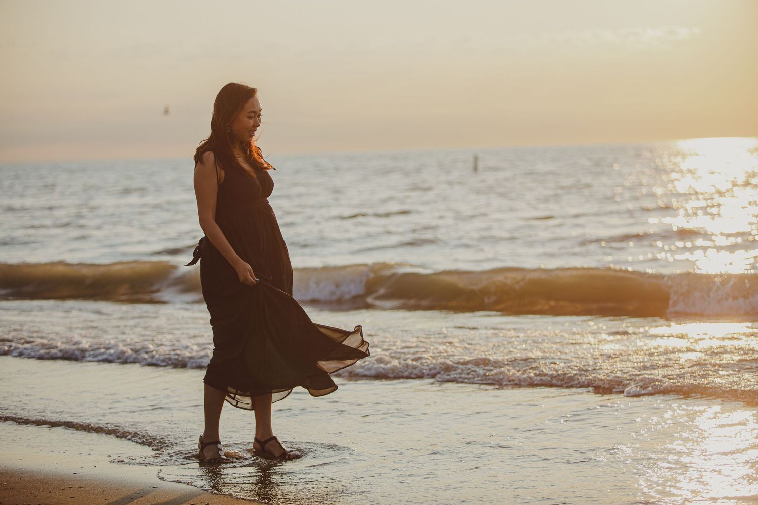Silhouetted figure in flowing dress stands at waters edge on beach during golden sunset.