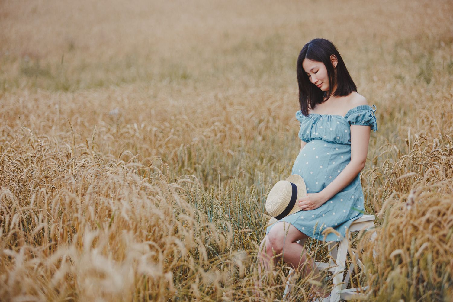 Someone in a light blue off-shoulder dress sits peacefully in a golden wheat field on a summer day.