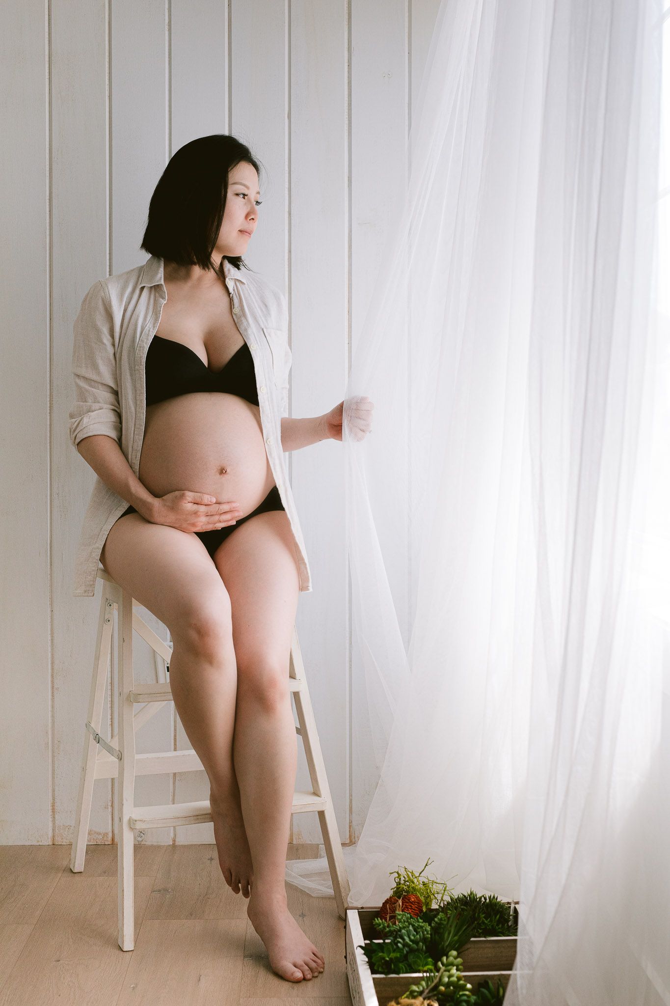 A minimalist maternity photo features someone in black lingerie and open shirt sitting on a white stool by sheer curtains.