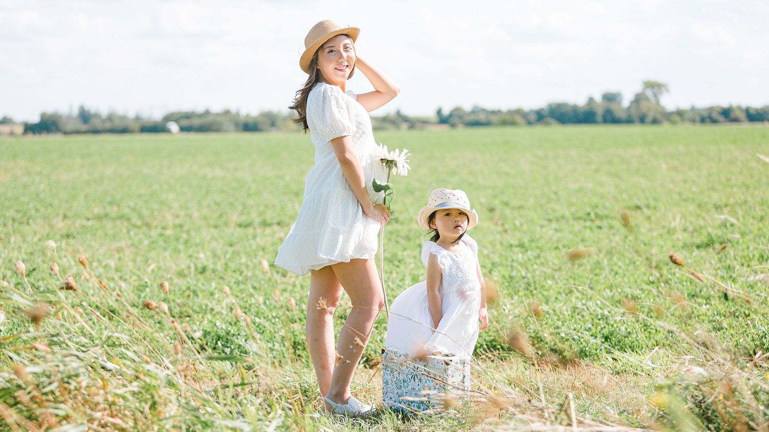 Two people in light summer dresses stand together in a sunlit meadow on a bright countryside day.