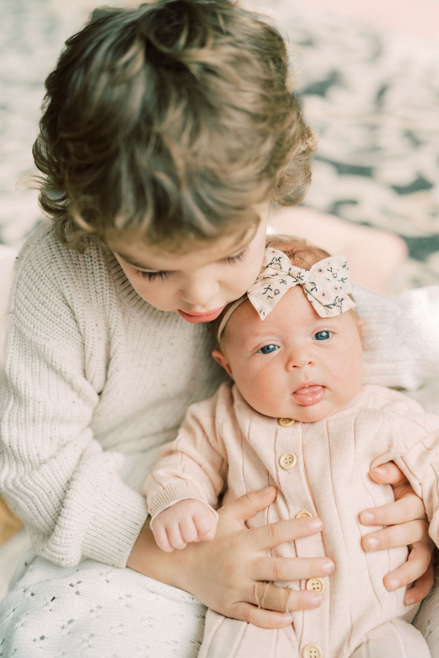 Siblings share intimate moment as older child holds infant wearing bow headband.