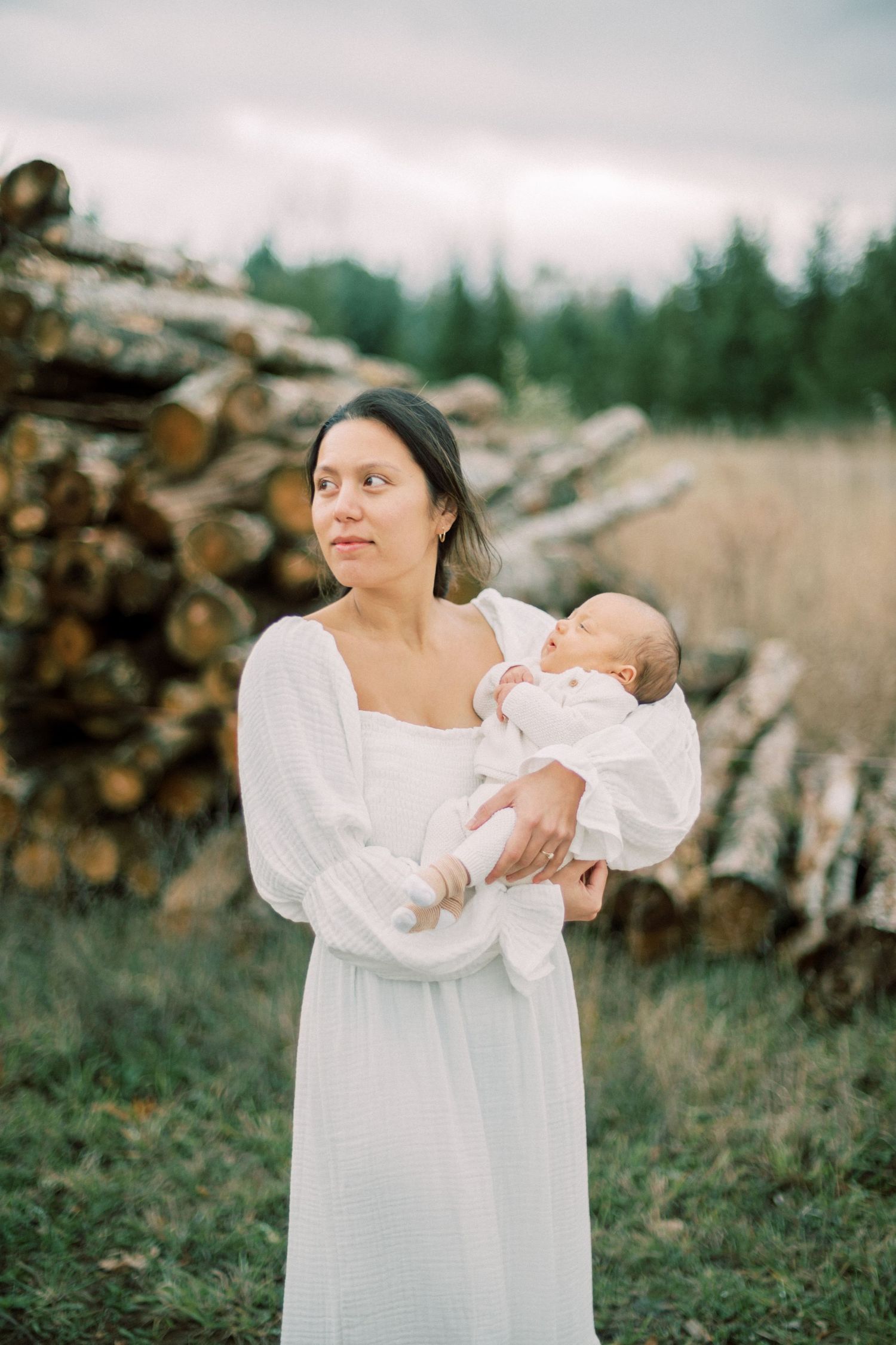 A woman in a white dress holds a baby against a backdrop of stacked firewood in a rural setting.