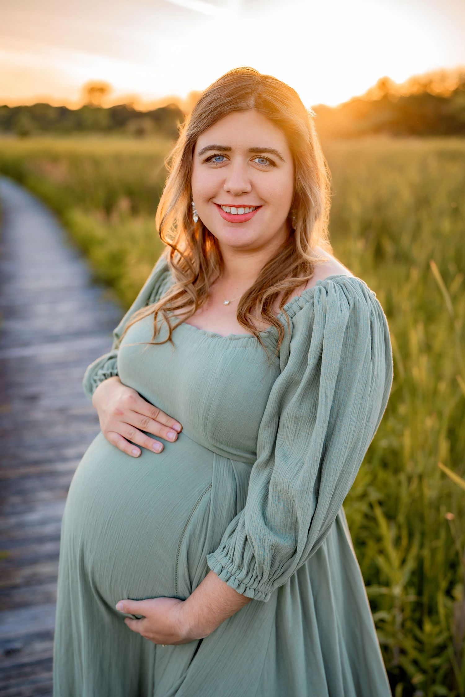 A woman in a sage green dress stands on a wooden boardwalk during golden hour, gently cradling her baby bump.