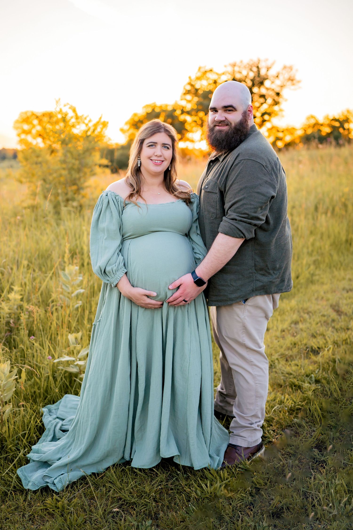 A couple poses in a golden field at sunset, with the woman wearing a flowing sage green maternity dress.
