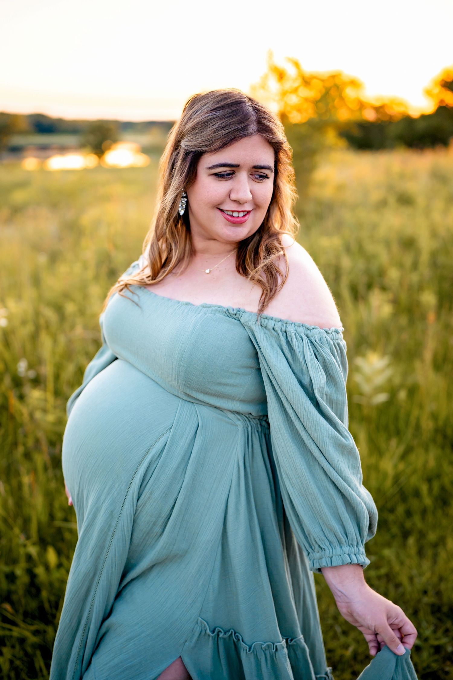 A pregnant woman in a sage green off-shoulder dress poses in a golden field at sunset.