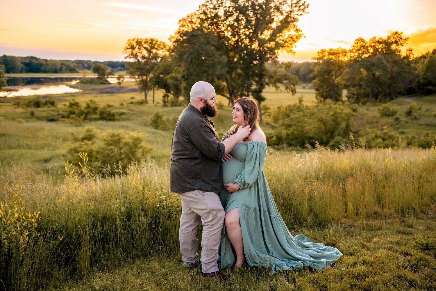 A couple embraces in a golden field at sunset with a pond and trees in the background.