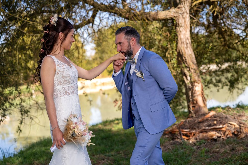 Couple in wedding attire posing together under a large tree near water.