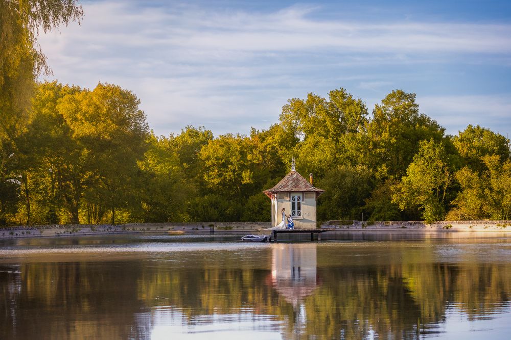 A small white gazebo reflects on a tranquil lake surrounded by golden autumn trees during sunset.