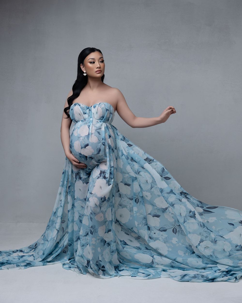 A flowing blue and white floral maternity gown with strapless sweetheart neckline and long train on grey background.