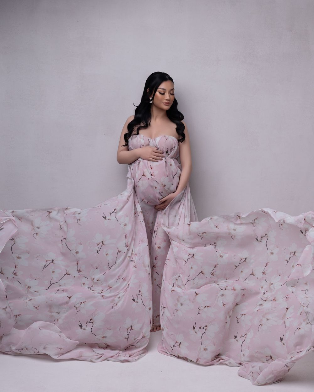 A pregnant woman in a pale pink floral gown poses elegantly against a gray backdrop with flowing fabric.