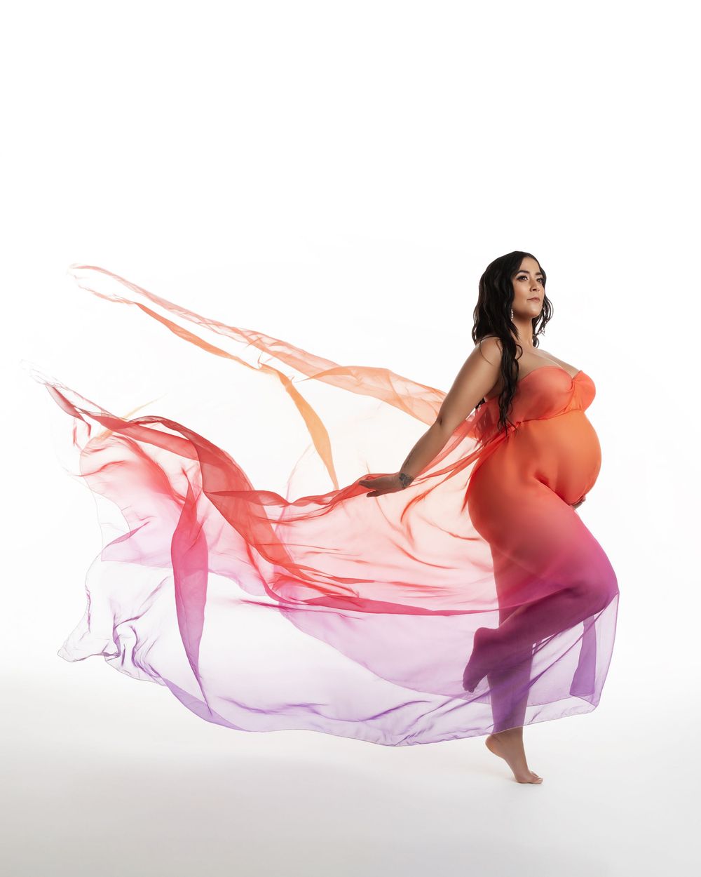 Woman in maternity dress with flowing coral and purple fabric creating ethereal movement against white backdrop.