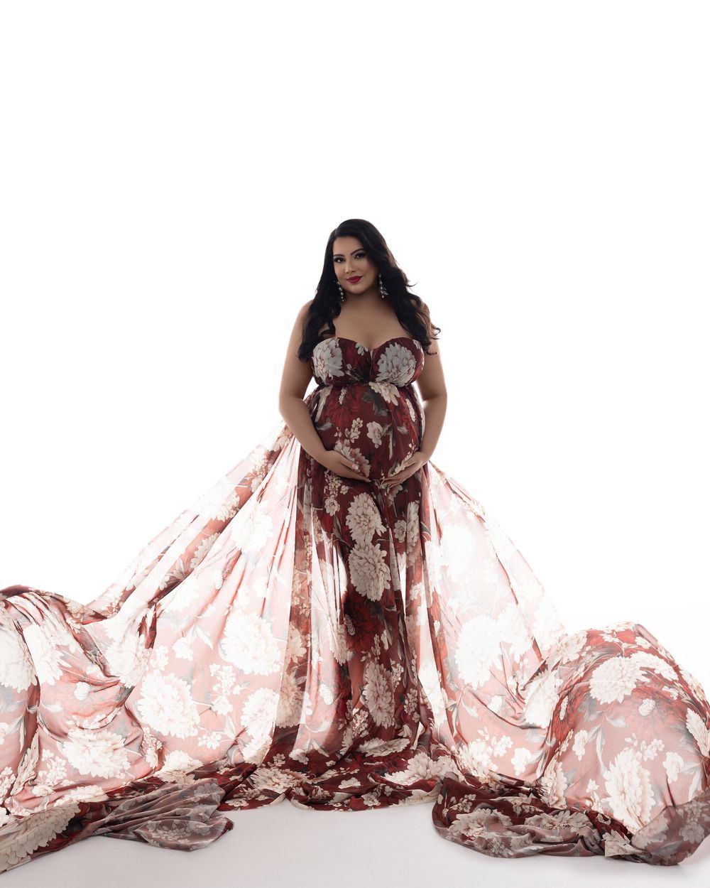A pregnant woman poses in a flowing brown and pink maternity gown with sweeping fabric against a white backdrop.