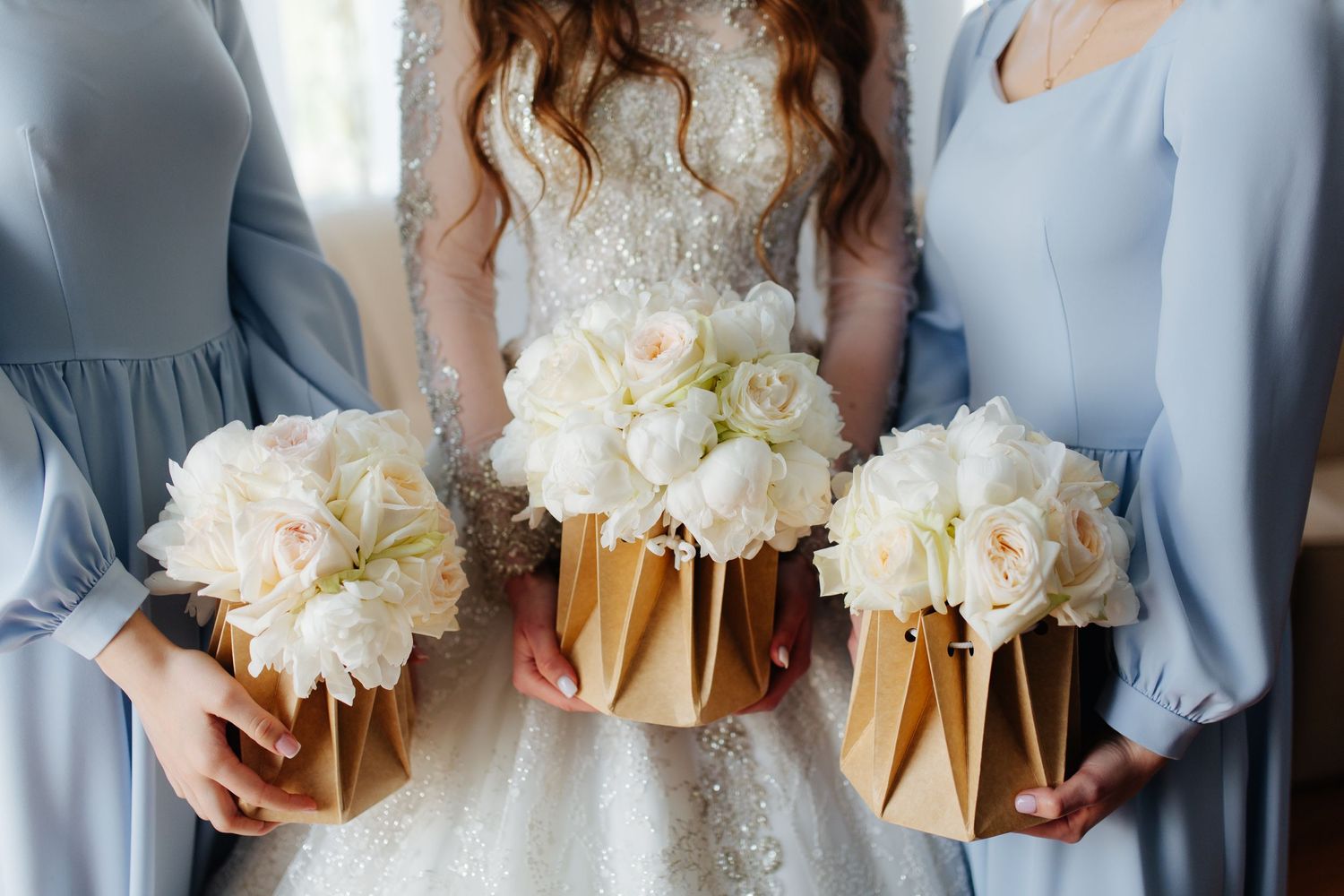 Wedding party holds elegant white rose bouquets wrapped in gold satin ribbon while wearing blue and lace dresses.