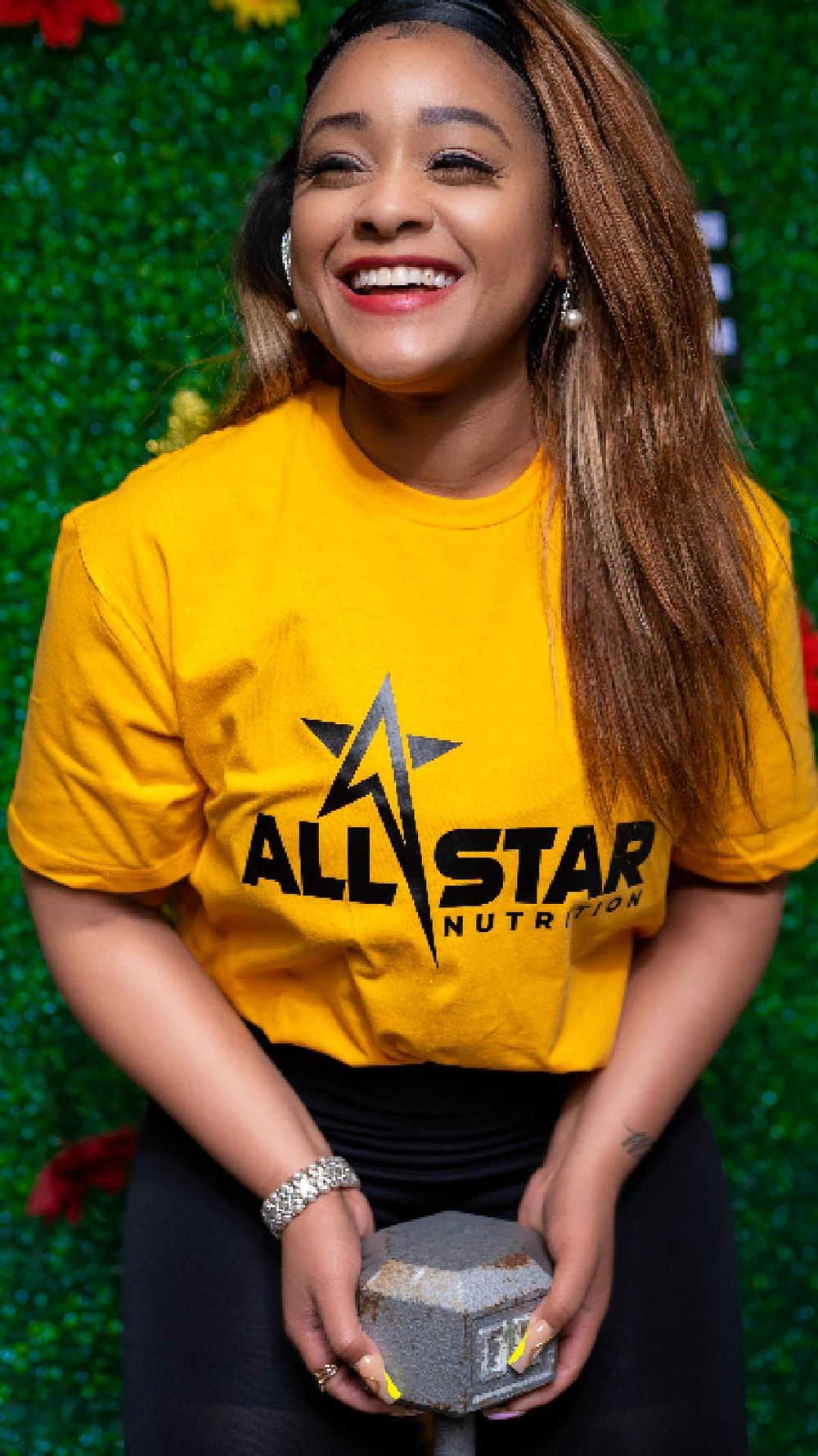 Person wearing yellow AllStar Nutrition t-shirt poses against green floral backdrop.