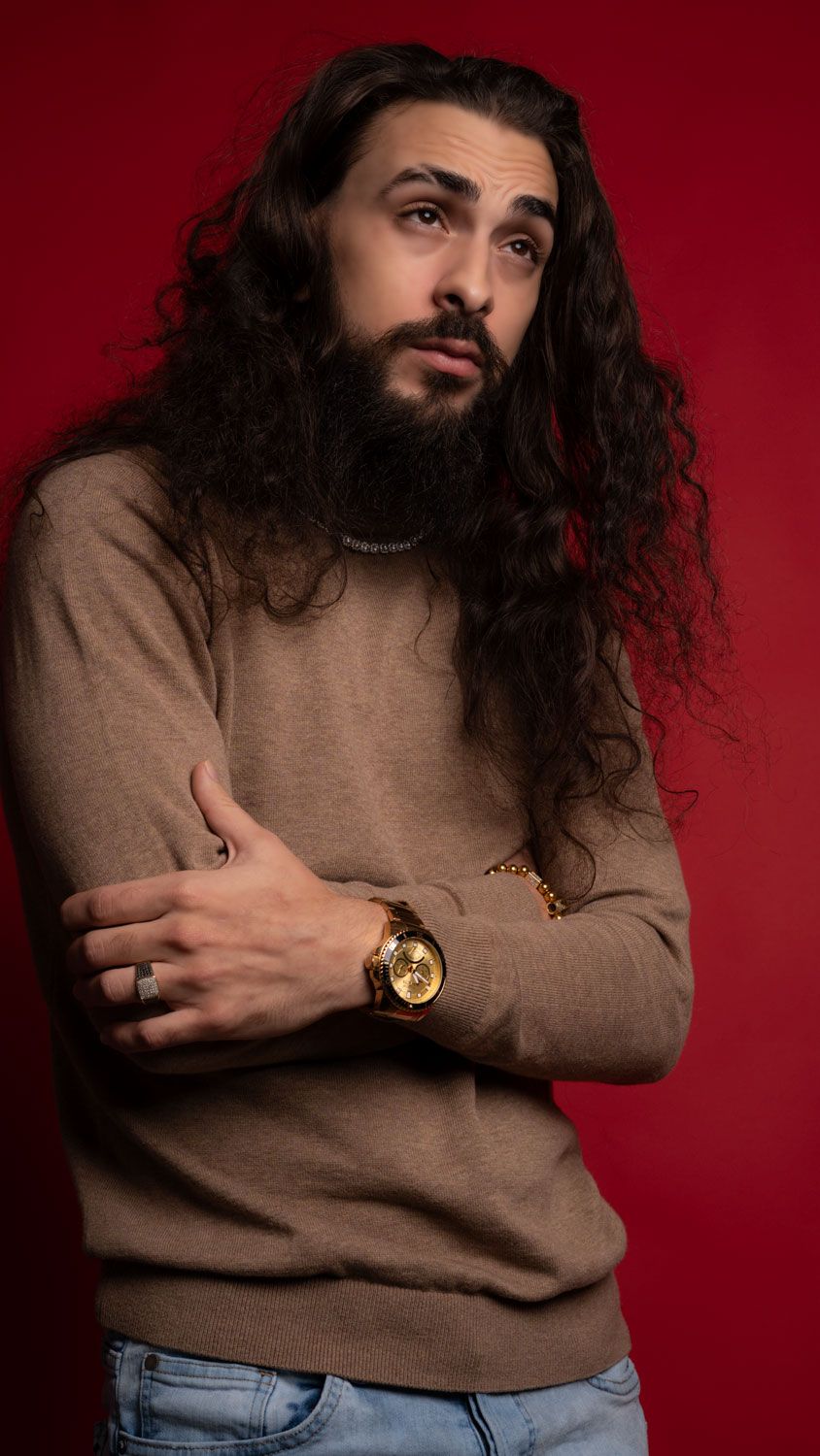 Portrait of man with long dark hair wearing beige sweater against red background.