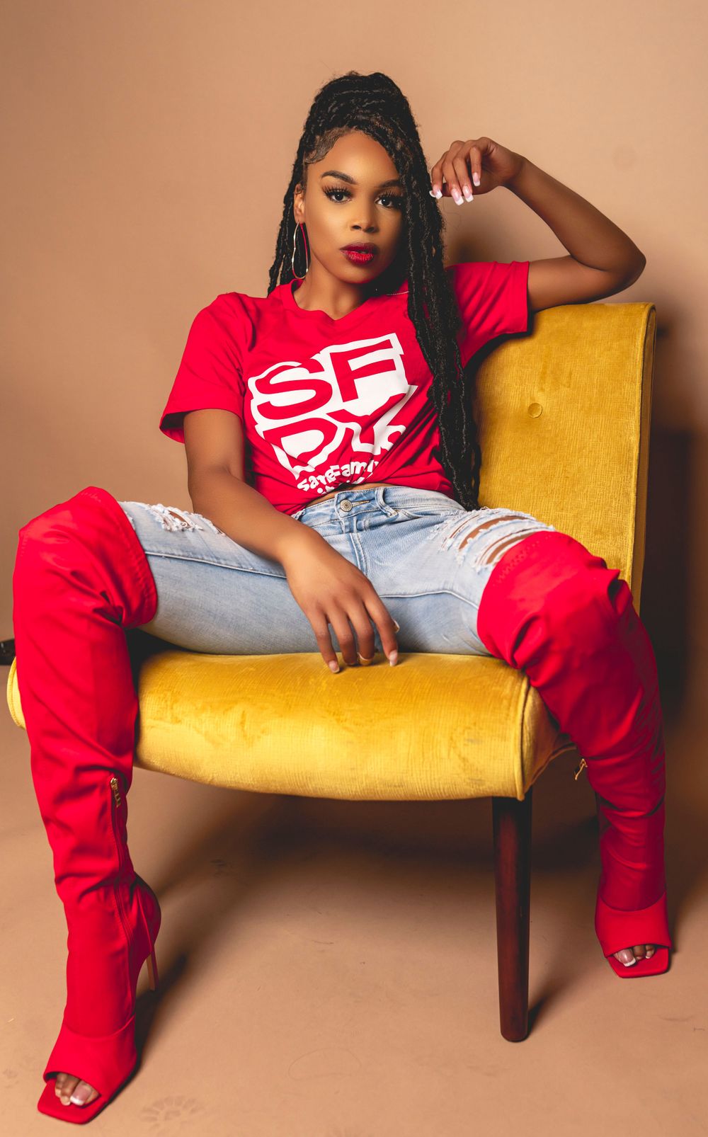 Model in red SF shirt and jeans poses on yellow velvet chair against beige background.